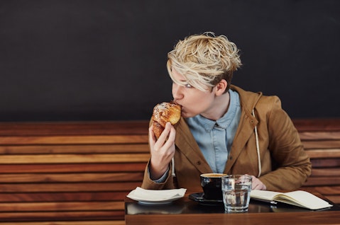 Young woman with trendy blonde hair looking away while taking a bite from her pastry, at a table in a modern cafe with a dark grey wall