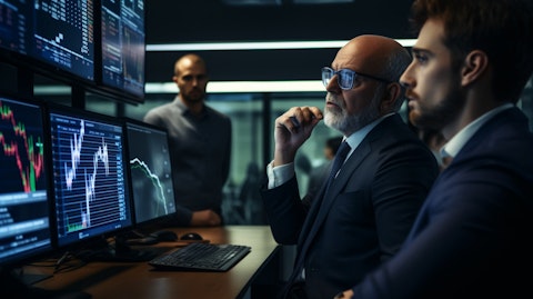A modern looking financial adviser sitting in front of a trading monitor, gesturing to a group of investors.
