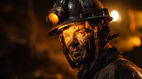A coal miner in a thick protective suit and helmet drilling for coal under bright lights.