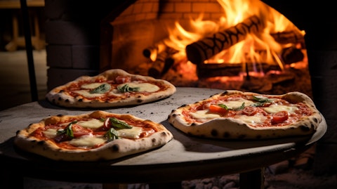 A stack of pizzas prepared in a wood-fired oven, with fresh ingredients laid out beside them in the kitchen.