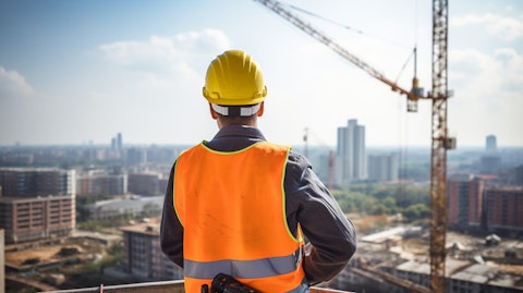 A worker in a safety helmet and bright orange vest surveying a construction site from a crane.