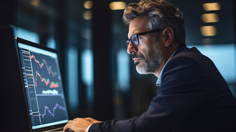 A close-up of a financial executive looking intently at their laptop screen, with a wall of financial charts in the background.
