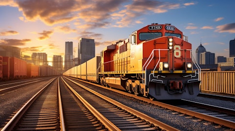 A freight train carrying railcar equipment in the foreground with a commercial area in the background.