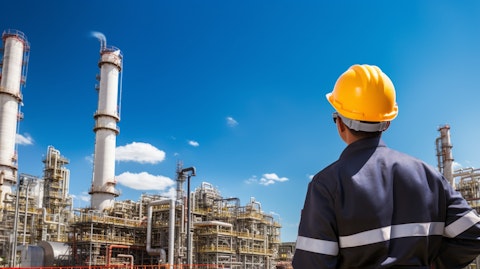 A worker in a hard hat standing in front of a giant oil refinery, the stark blue sky and grey refinery in the background.