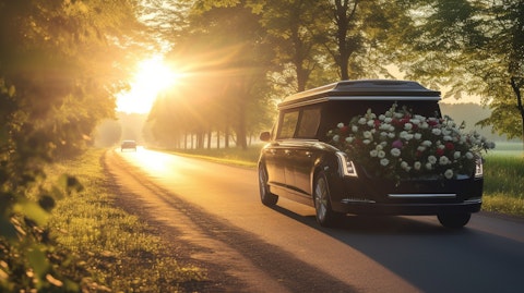 A funeral procession travelling along a rural road with a hearse pulling a casket covered in flowers.