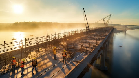 An aerial view of a bridge under construction with workers continuing their work despite the early morning light.