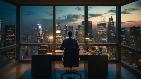 A business man sitting at a desk in a high rise office, looking out the window at the financial district below.